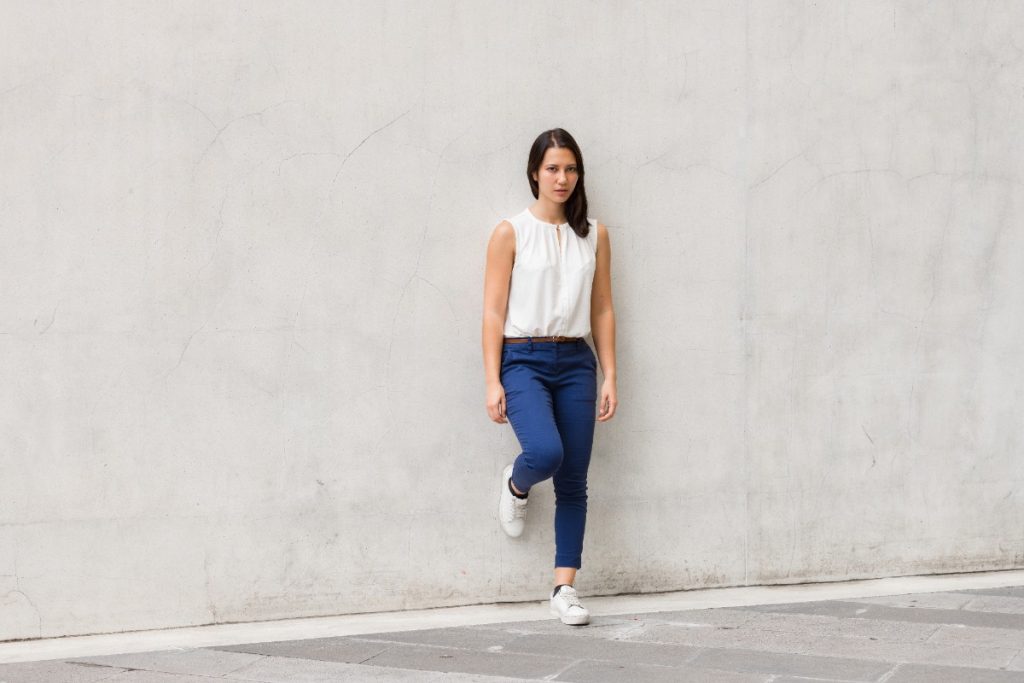 Une femme posant contre le mur en portant un haut blanc, un pantalon bleu avec un basket